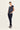 A woman modeling a navy blue short-sleeve top with white vertical stripes and dark navy pants by Verge, standing against a plain off-white studio background.
