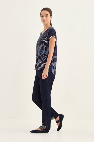 A woman modeling a navy blue short-sleeve top with a white and light blue geometric pattern, paired with dark navy pants by Verge and black flats, against a plain off-white studio background.