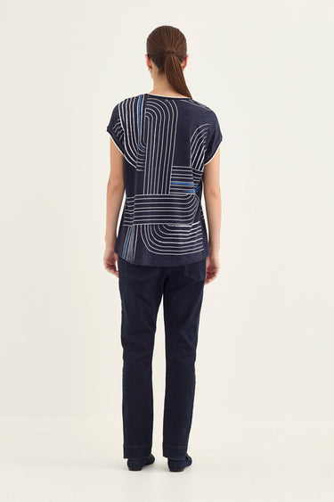 A woman viewed from the back wearing a navy blue short-sleeve top with a white geometric line pattern and dark navy pants by Verge, standing against a plain off-white studio background.
