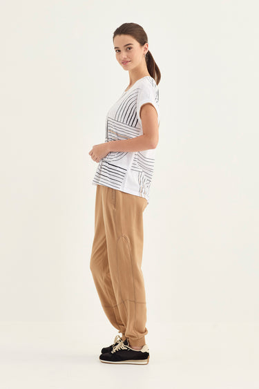 A woman modeling a white short-sleeve t-shirt with black diagonal stripes and beige wide-leg pants by Verge, standing against a plain white studio background.