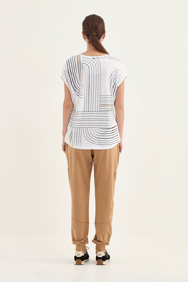 A woman viewed from behind wearing a white short-sleeve top with a black geometric stripe pattern, tan pants by Verge, and black sneakers, standing against a plain light background.