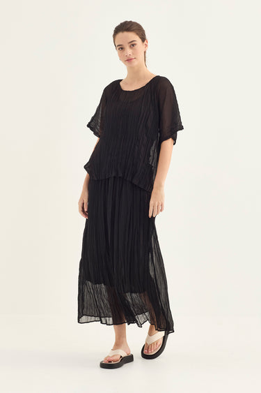 A woman modeling a black sheer, layered midi dress with flutter sleeves and a pleated skirt by Verge, paired with black slide sandals, against a plain off-white studio background.
