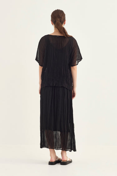 A woman viewed from behind wearing a black sheer pleated short-sleeve top and matching pleated maxi skirt by Verge, standing against a plain light background.