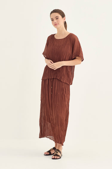 A woman modeling a rust-brown pleated two-piece set consisting of a short-sleeve top and a matching maxi skirt by Verge, paired with black slide sandals, against a plain off-white studio background.