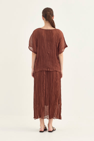 A woman viewed from behind wearing a rust-brown pleated two-piece outfit with a short-sleeve top and a long pleated skirt by Verge, paired with black sandals, against a plain off-white studio background.
