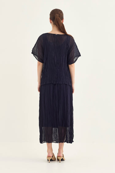 A woman viewed from the back wearing a dark navy pleated top and skirt with short flutter sleeves by Vergeand beige pointed-toe heels, against a plain light background.