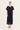 A woman modeling a dark navy blue pleated midi dress with short flutter sleeves and a buttoned waist by Verge, paired with beige flat sandals, against a plain off-white studio background.