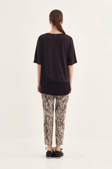 A woman viewed from behind wearing a loose black short-sleeve top, beige and brown snakeskin print pants by Verge, and black slip-on shoes, set against a plain off-white studio background.