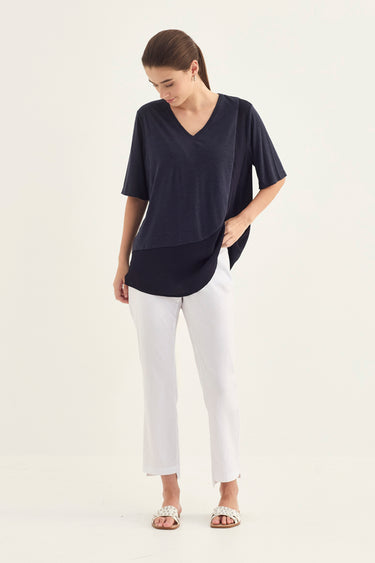 A woman modeling a navy blue short-sleeve V-neck top with white cropped pants by Verge and flat sandals, standing against a plain off-white studio background.