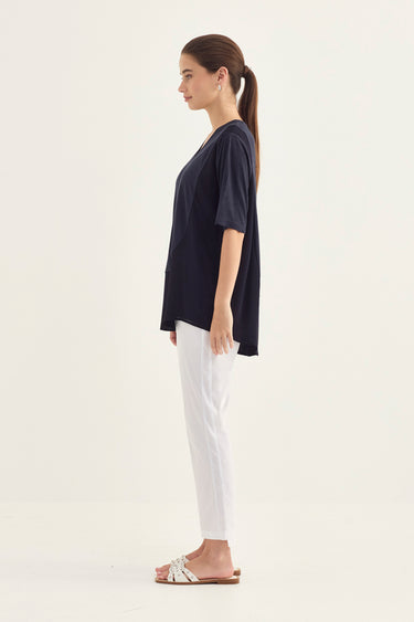 A woman modeling a navy blue short-sleeve V-neck top with white pants by Verge and flat sandals, standing against a plain off-white studio background.