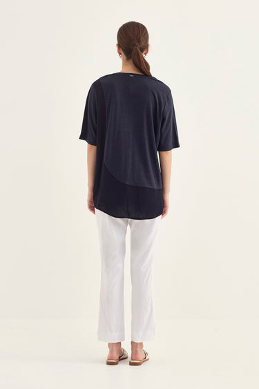 A woman viewed from behind wearing a navy blue short-sleeve V-neck top with white pants by Verge and flat sandals, standing against a plain off-white studio background.
