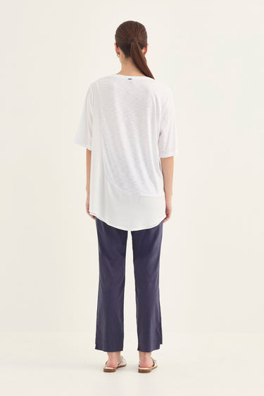 A woman viewed from behind wearing a loose white short-sleeve top and dark navy wide-leg pants by Verge, standing against a plain off-white studio background.