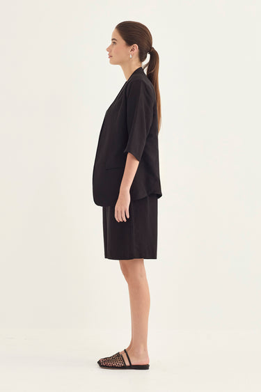 A woman in a side profile wearing a black short-sleeve blazer and matching black shorts by Verge, paired with black strappy flat sandals, against a plain off-white studio background.