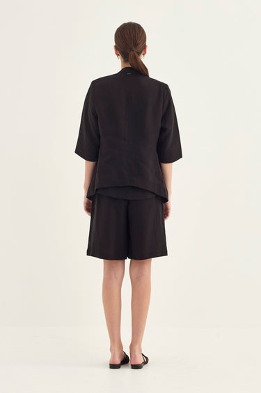 A model viewed from the back wearing a black short-sleeve cropped jacket and matching black shorts by Verge against a plain off-white studio background.