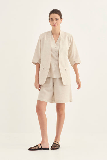 A woman modeling a light beige linen two-piece suit consisting of a short-sleeve button-front blazer and matching shorts by Verge, paired with black woven flat sandals, against a plain white studio background.