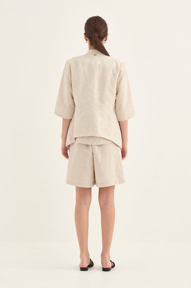 A model viewed from the back wearing a light beige linen two-piece set consisting of a short-sleeve cropped jacket and matching shorts by Verge, paired with black flat sandals against a plain off-white studio background.
