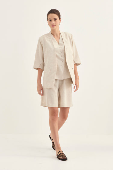 A woman modeling a light beige linen two-piece suit consisting of a short-sleeve button-front vest and matching shorts by Verge, paired with black patterned flat sandals, against a plain white studio background.