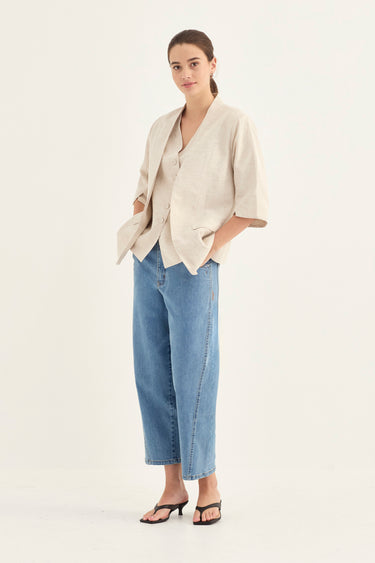 A woman modeling a light beige linen short-sleeve blazer and wide-leg blue jeans by Verge against a plain off-white studio background.