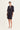 A woman modeling a dark navy blue tailored set consisting of a short-sleeve blazer and matching skirt by Verge, worn over a black and white striped top with white slide sandals, against a plain light background.