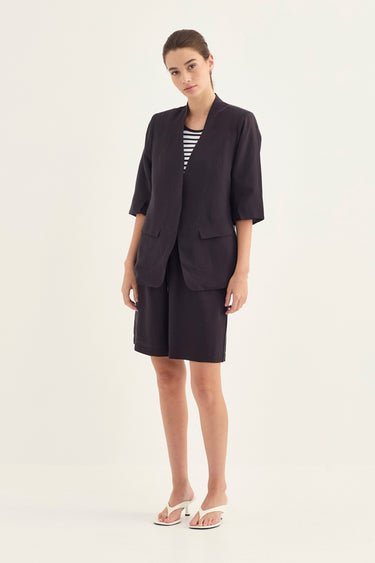 A woman modeling a dark navy blue tailored set consisting of a short-sleeve blazer and matching skirt, worn over a black and white striped top with white slide sandals by Verge, against a plain light background.