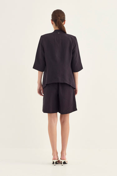 A model viewed from the back wearing a dark charcoal gray short-sleeve top and matching shorts by Verge, paired with white and black pointed-toe heels, against a plain off-white studio background.