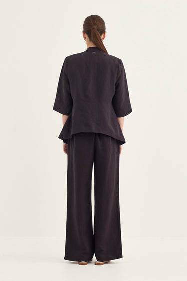 A model viewed from the back wearing a dark plum-colored two-piece linen set consisting of a short-sleeve peplum-style top and matching wide-leg trousers by Verge, set against a plain off-white studio background.