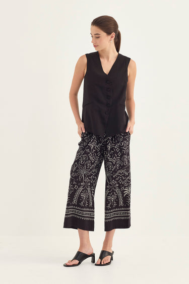 A woman modeling a sleeveless black vest and black cropped pants with a white paisley pattern by Verge, paired with black slide sandals, against a plain off-white studio background.