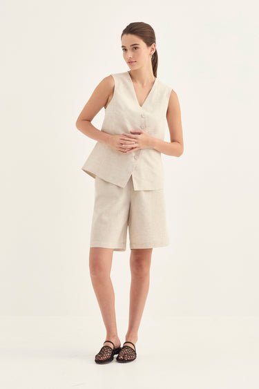 A woman modeling a light beige linen two-piece set consisting of a sleeveless V-neck vest and matching shorts by Verge, paired with black lace-up sandals, against a plain white studio background.