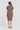 A woman viewed from the back wearing a short-sleeve, knee-length textured dress in a muted cocoa color by Verge, paired with black flat sandals, against a plain light gray studio background.