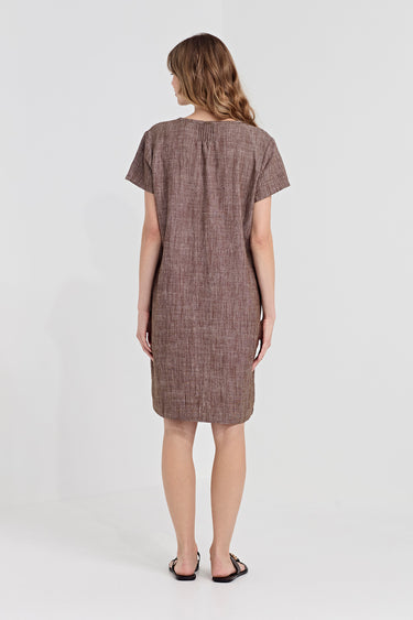 A woman viewed from the back wearing a short-sleeve, knee-length textured dress in a muted cocoa color by Verge, paired with black flat sandals, against a plain light gray studio background.