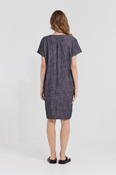 A woman viewed from the back wearing a short-sleeve knee-length dress with a textured by Verge, heathered dark gray and purple fabric. She is standing against a plain light gray studio background.