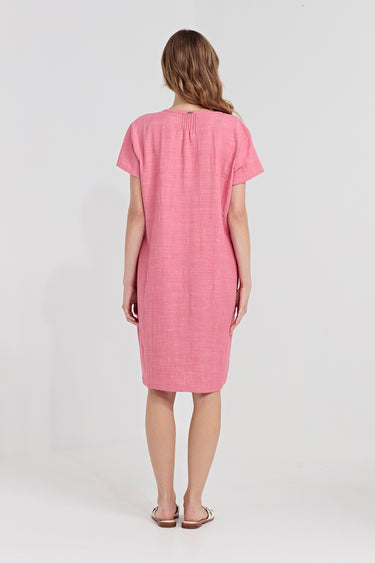 A woman viewed from behind wearing a short-sleeve pink linen dress by Verge with a gathered neckline, standing against a plain white studio background.