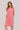 A woman modeling a short-sleeve pink linen dress by Verge with a split neckline and side pockets, set against a solid turquoise background.