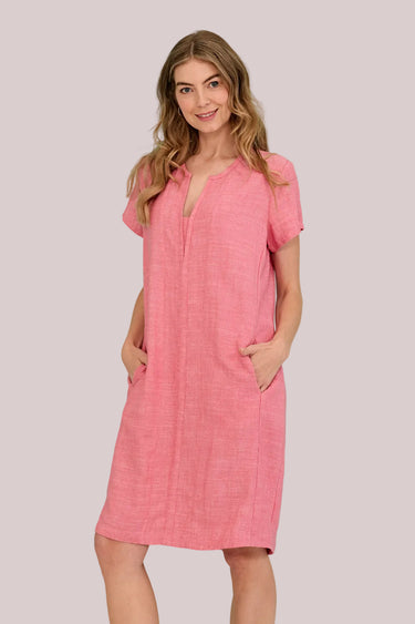 A woman modeling a short-sleeve pink linen dress by Verge with a split neckline and side pockets, set against a solid turquoise background.