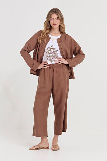A woman modeling a matching brown two-piece lounge set consisting of a cropped jacket and wide-leg cropped pants, worn over a white graphic t-shirt by Verge and flat sandals, against a light gray studio background.