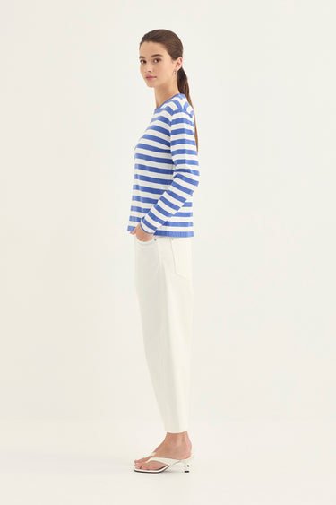 A woman stands in profile wearing a blue and white horizontally striped long-sleeve top, white pants by Verge, and white heeled sandals against a plain light background.