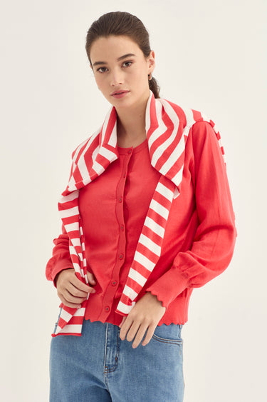 A woman wearing a red long-sleeve button-up shirt with a large white and red striped draped collar, paired with blue jeans by Verge, against a plain light background.