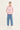 A woman modeling a pink and white horizontally striped long-sleeve knit top with light blue wide-leg jeans by Verge and white sneakers, standing against a plain light gray studio background.