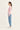 A woman in profile wearing a pink and white striped long-sleeve top, light blue jeans by Verge, and white sneakers, standing against a plain off-white studio background.