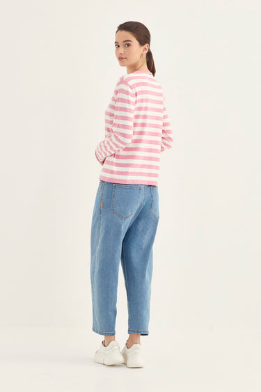 A woman shown from the back wearing a pink and white striped long-sleeve top, light blue cropped jeans by Verge, and white sneakers, standing against a plain off-white studio background.