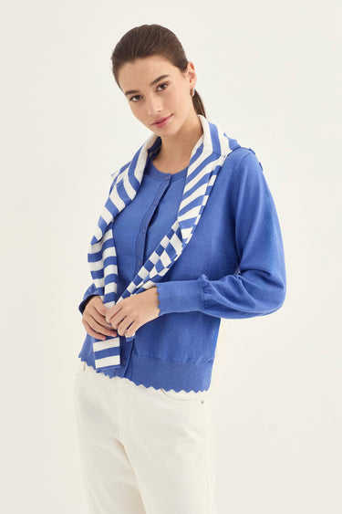 A woman modeling a blue knit cardigan with a scalloped hem and a blue-and-white striped knit sweater by Verge over her shoulders, paired with white pants, against a plain light background.