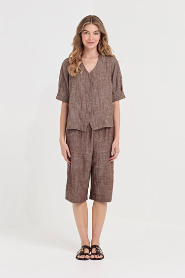 A woman modeling a matching brown linen two-piece set consisting of a short-sleeve button-front shirt and matching knee-length shorts by Verge, paired with black sandals, against a plain white studio background.