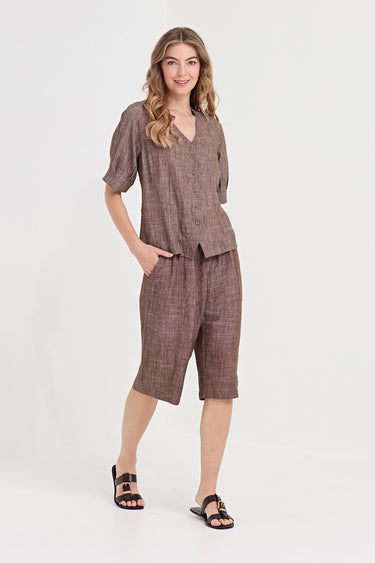 A woman modeling a matching two-piece linen set consisting of a short-sleeve button-front top and matching cropped shorts by Verge in a brown and beige textured weave, paired with black slide sandals, against a plain white studio background.