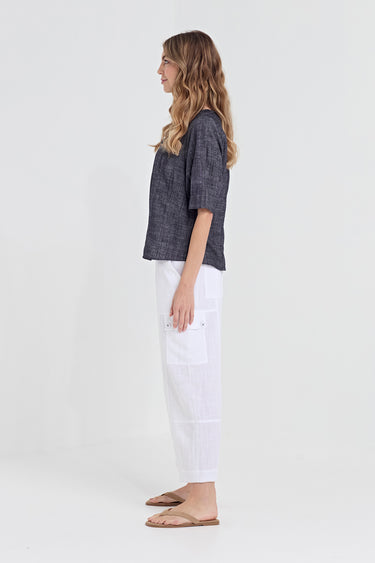 A woman stands in profile wearing a dark charcoal gray textured short-sleeve top, white wide-leg pants by Verge, and tan flat sandals against a plain white studio background.