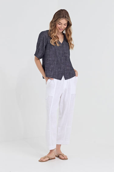 A woman modeling a short-sleeve textured dark gray top and white cropped pants with side pockets by Verge, paired with tan sandals, against a plain light gray studio background.