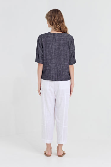 A woman viewed from the back wearing a dark textured short-sleeve top and white cropped pants by Verge, standing against a plain light gray studio background.