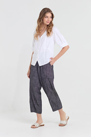 A woman modeling a white short-sleeve button-up shirt and dark gray wide-leg cropped pants by Verge with white slide sandals, photographed against a light gray studio background.