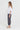 A woman stands in profile wearing a loose white short-sleeve top, dark charcoal plaid cropped pants by Verge, and white slide sandals against a plain light gray studio background.