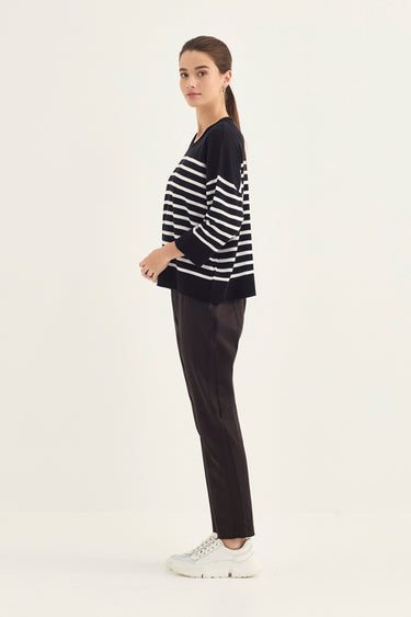 A woman stands in profile wearing a black and white horizontally striped long-sleeve knit top, dark brown trousers by Verge, and white sneakers against a plain white studio background.