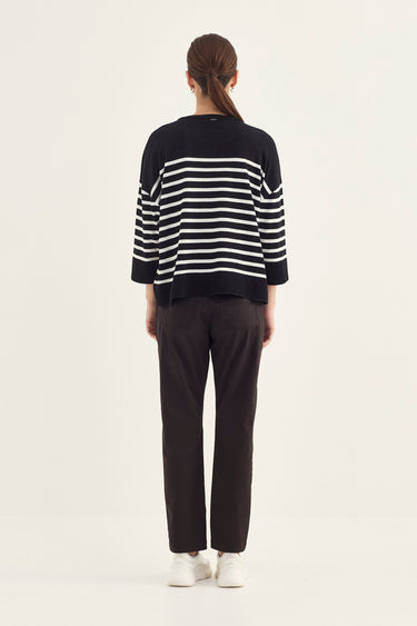 A woman viewed from behind wearing a black knit sweater with white horizontal stripes on the sleeves and upper back, paired with dark brown pants by Verge and white sneakers, against a plain off-white studio background.